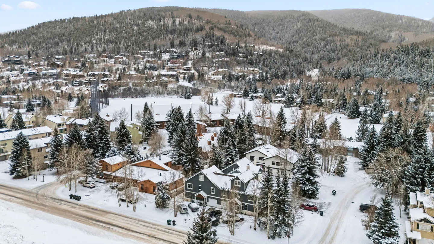 Aerial view of a snowy mountain neighborhood featuring homes surrounded by snow-covered pine trees and hills.