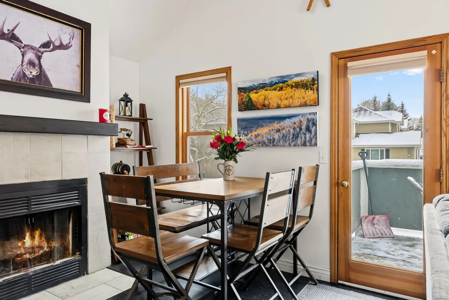 Dining nook with a wooden table, four metal and wood chairs, a lit fireplace, and an exterior glass door leading to a snowy balcony.