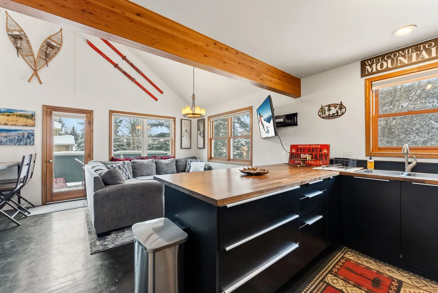 Open-concept kitchen and living area with black cabinets, butcher block countertops, exposed wooden ceiling beam, and ski decor.