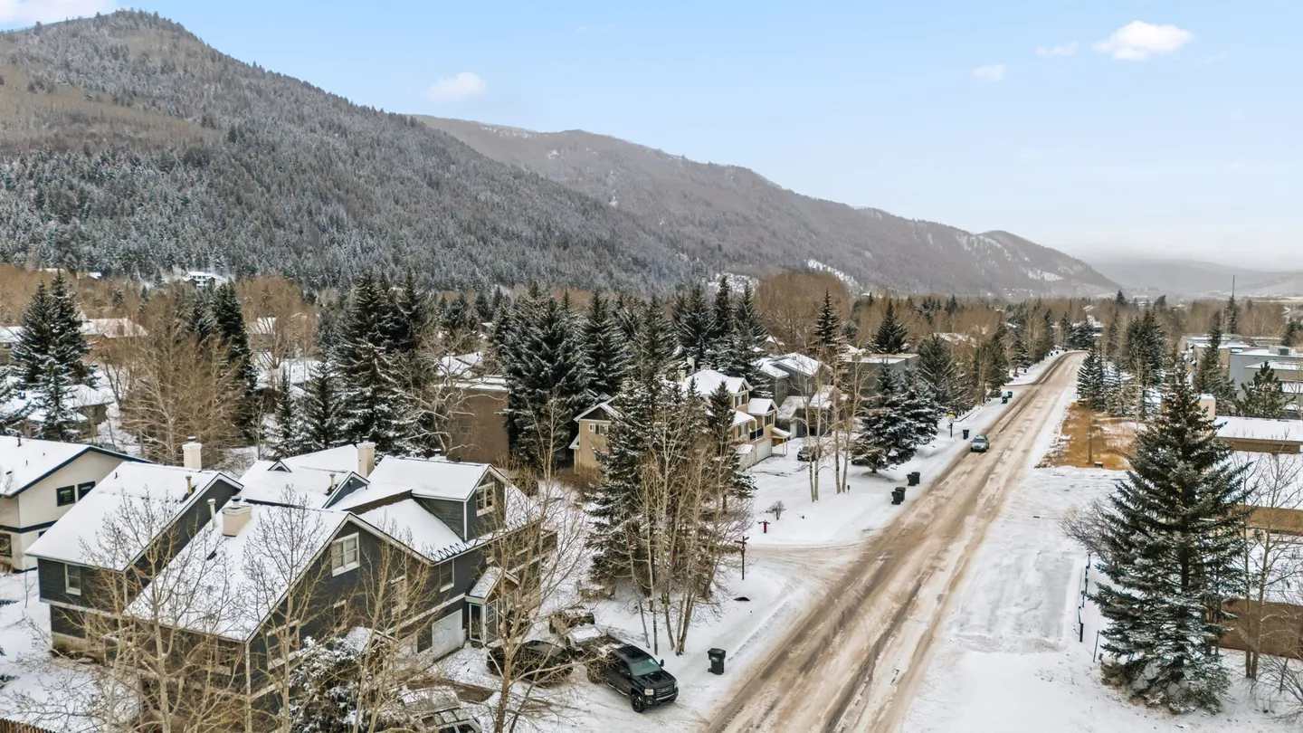 Elevated view of a snow-covered residential street and houses backed by a forested mountain ridge.