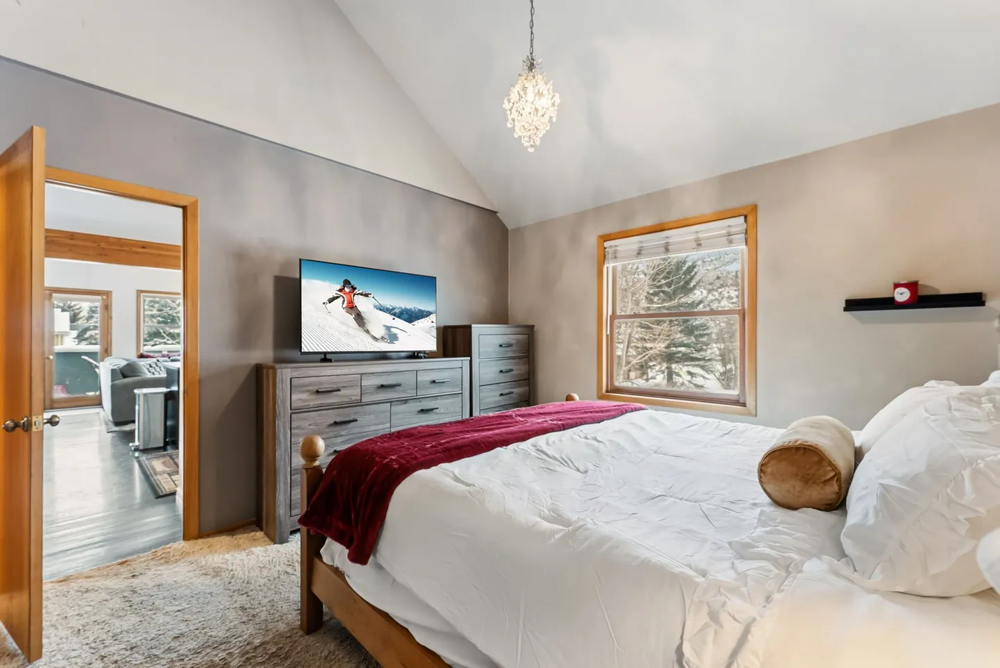 Bedroom featuring vaulted ceilings, a chandelier, carpet flooring, a bed with white and red bedding, and a wall-mounted TV on a wooden dresser.