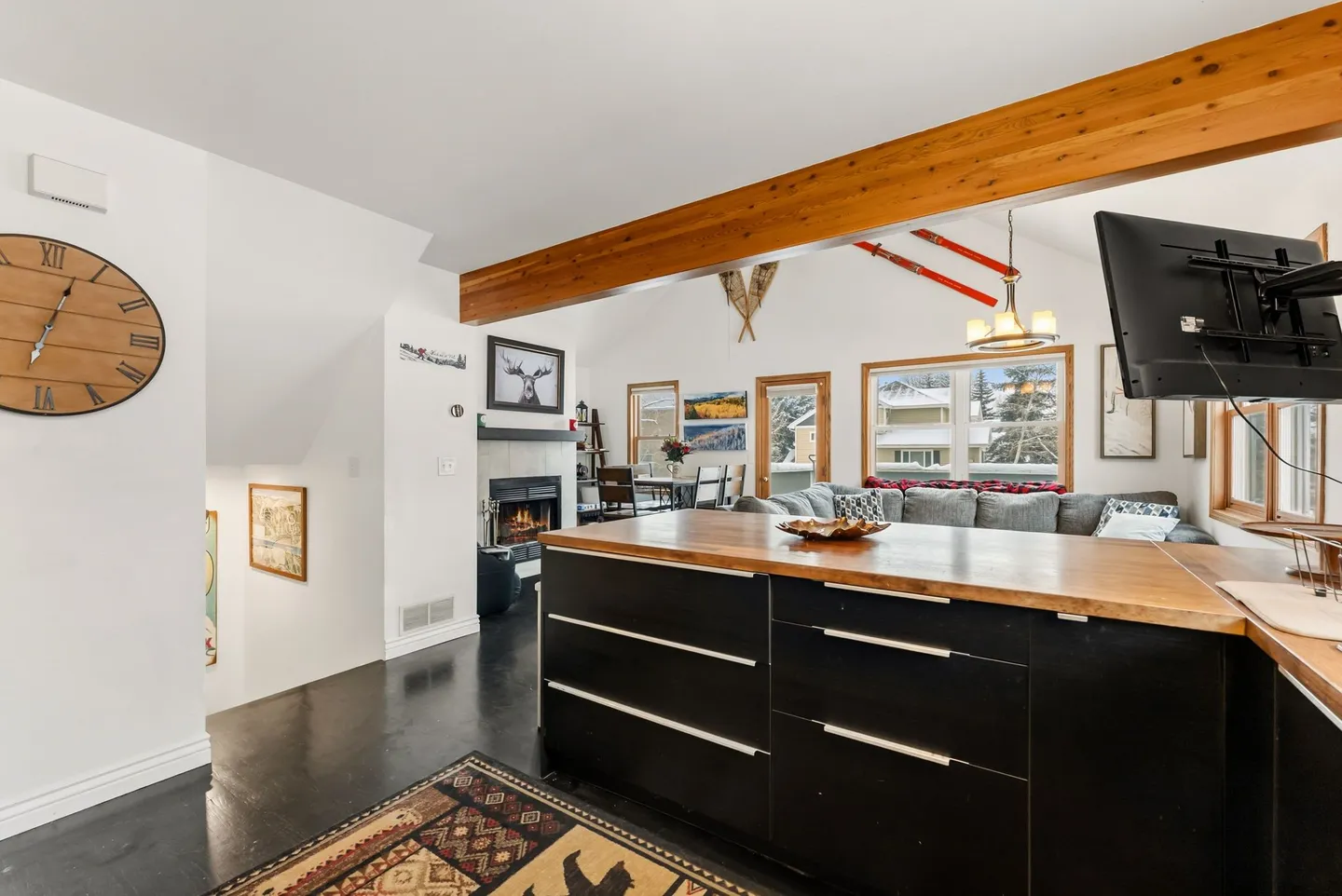 Open-concept living space viewed from a kitchen island, featuring a fireplace, seating area, and vaulted ceilings with exposed wood beams.