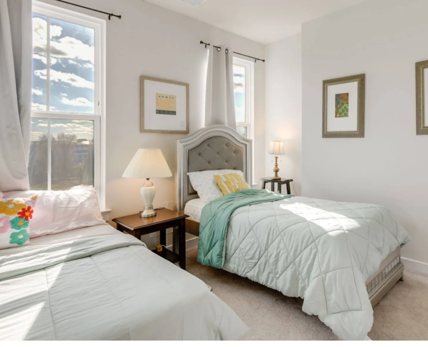 Bright bedroom with two twin beds, bedside tables with lamps, and large windows showing a blue sky.