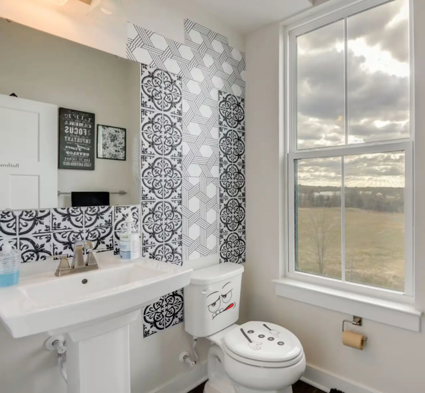 Bathroom with white pedestal sink, toilet decorated with cartoon face stickers, patterned black and white wall tiles, and a window overlooking a field.