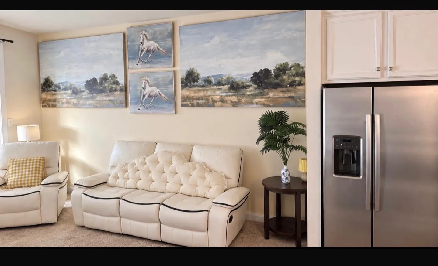 Living area with white leather seating, landscape and horse artwork, and a partial view of a stainless-steel refrigerator.