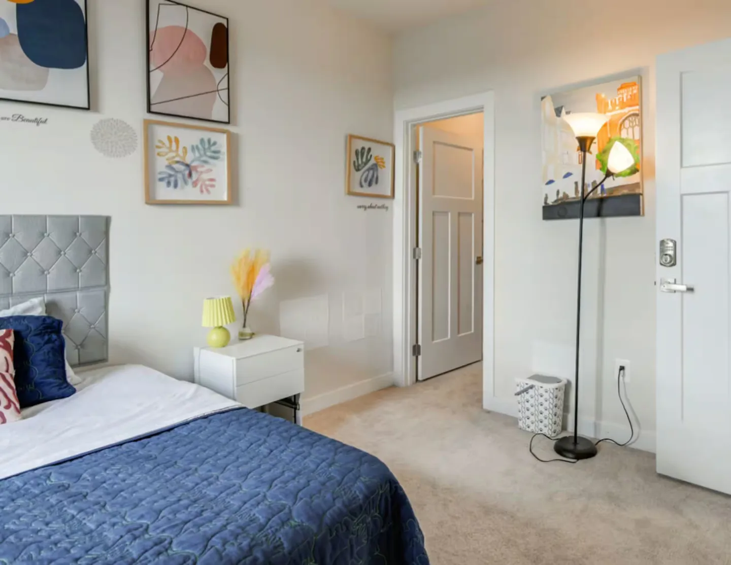Bedroom with blue bedding, a silver tufted headboard, carpeted flooring, wall art, and an open doorway.