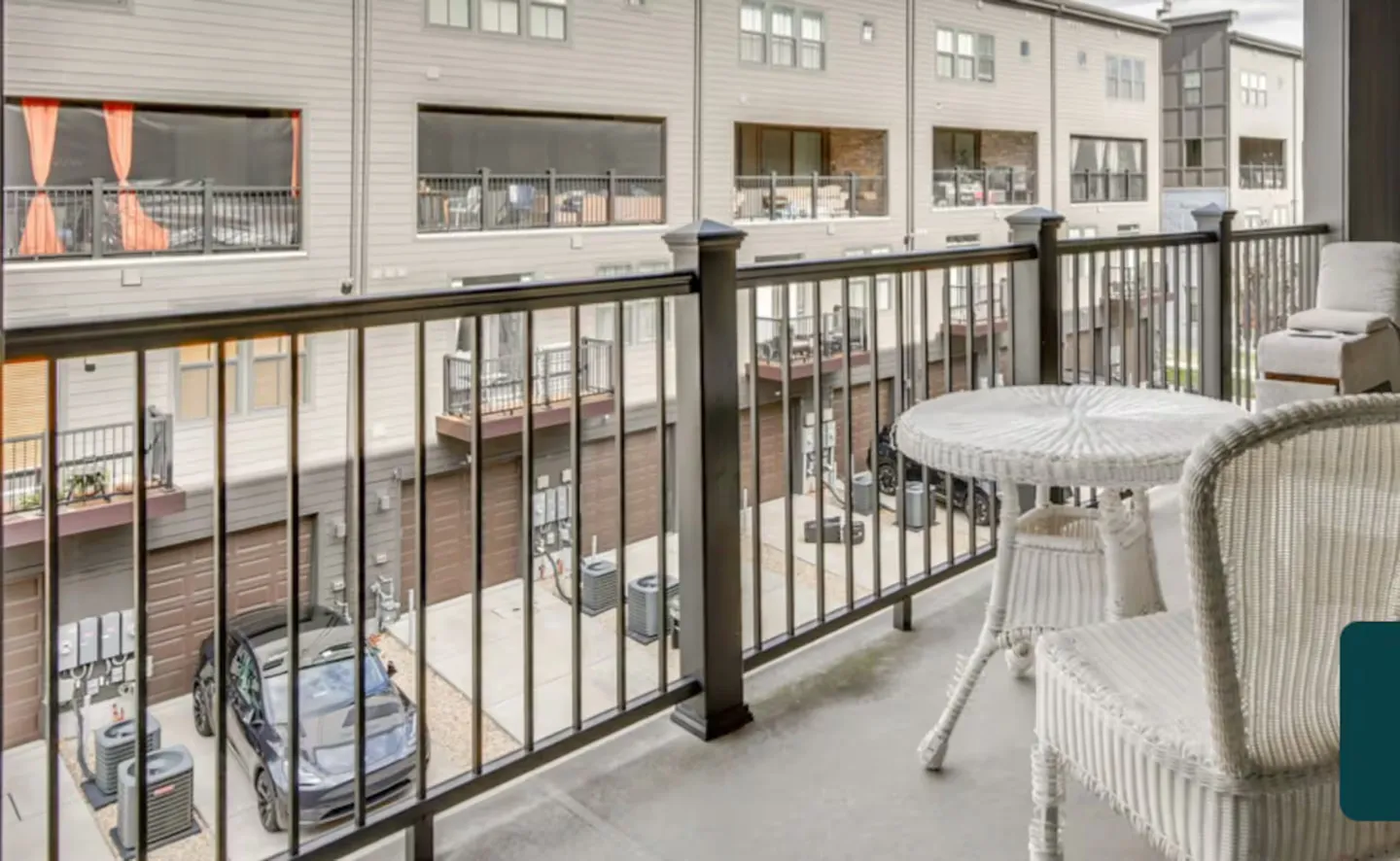 Balcony with black metal railings, a white wicker chair, and a small round table overlooking adjacent townhomes.