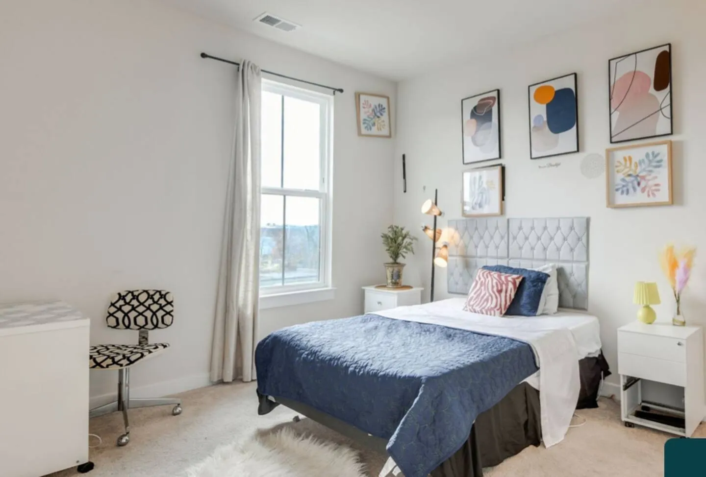 Bedroom with a bed dressed in blue and white, framed abstract art on the wall, a window, and a small desk with an office chair.