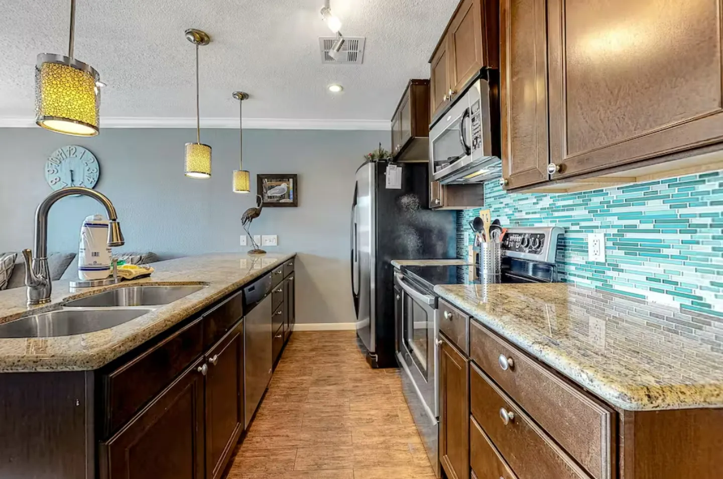 A kitchen with dark wood cabinets, speckled granite countertops, stainless steel appliances, and a turquoise tile backsplash.