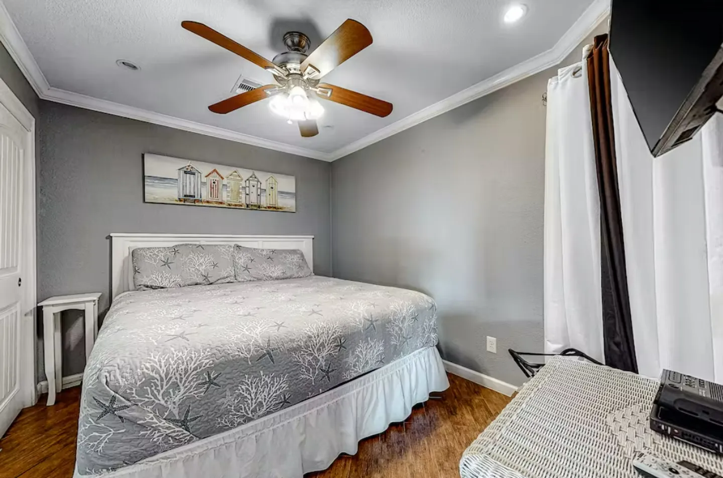 Bedroom with a white headboard, coastal-patterned bedspread, wood-look flooring, ceiling fan, and framed beach hut wall art.