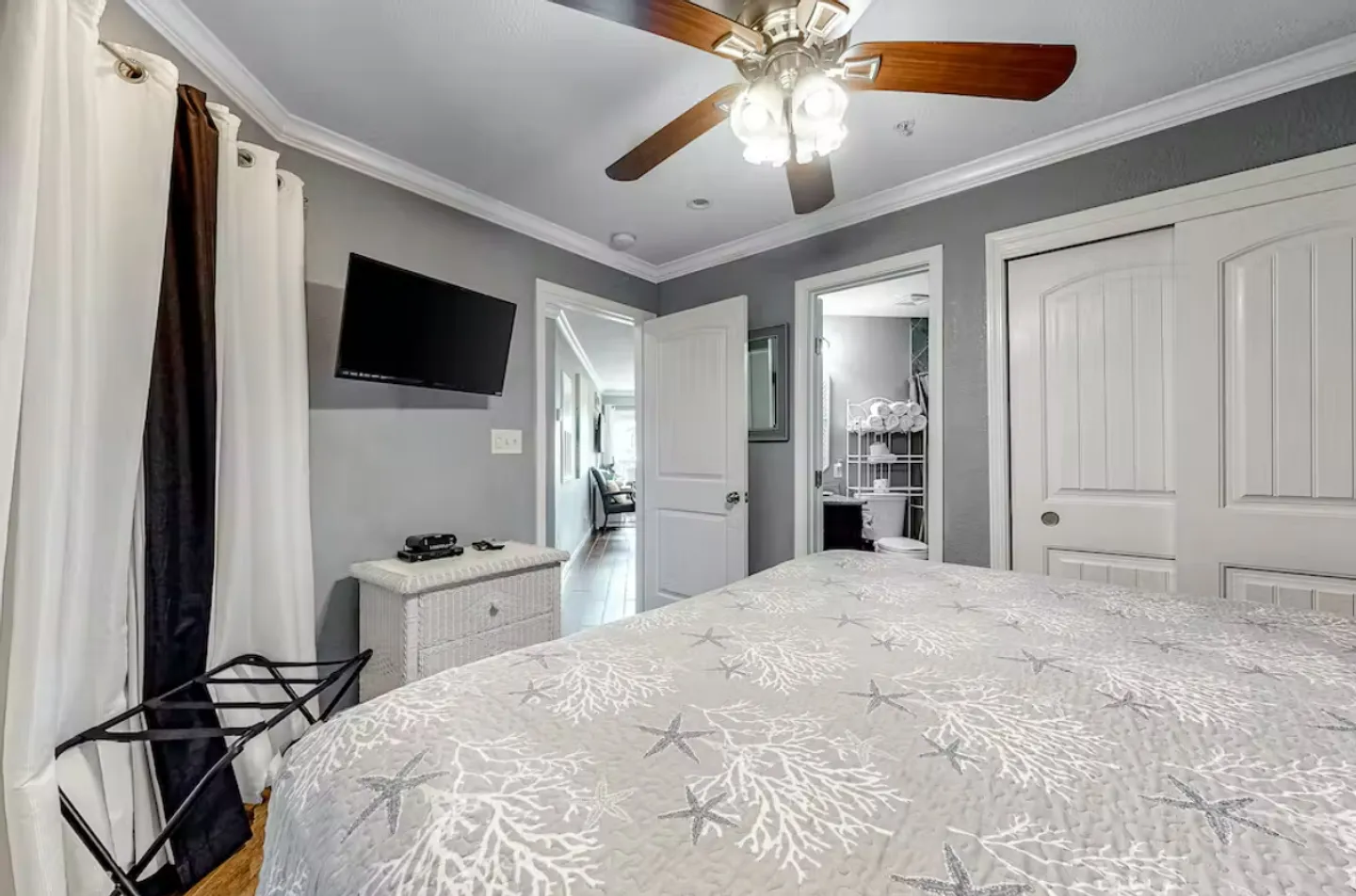 Bedroom featuring a bed with starfish-patterned bedding, a ceiling fan, a wall-mounted TV, and an open door to an en-suite bathroom.