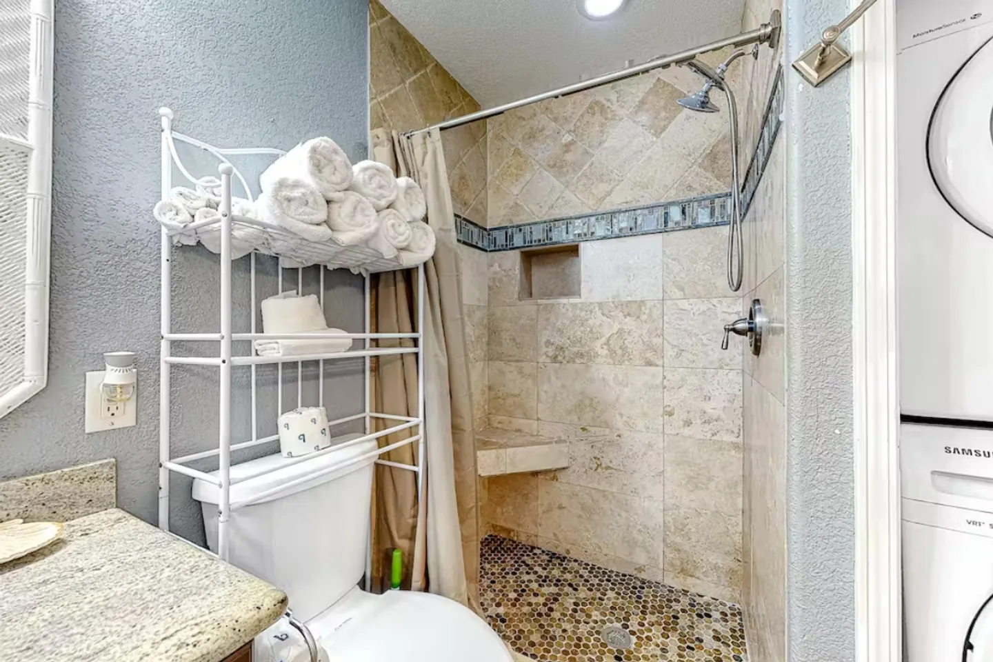 Bathroom showing a tiled walk-in shower with pebble floor, toilet with over-the-toilet wire shelving, and stacked laundry appliances.