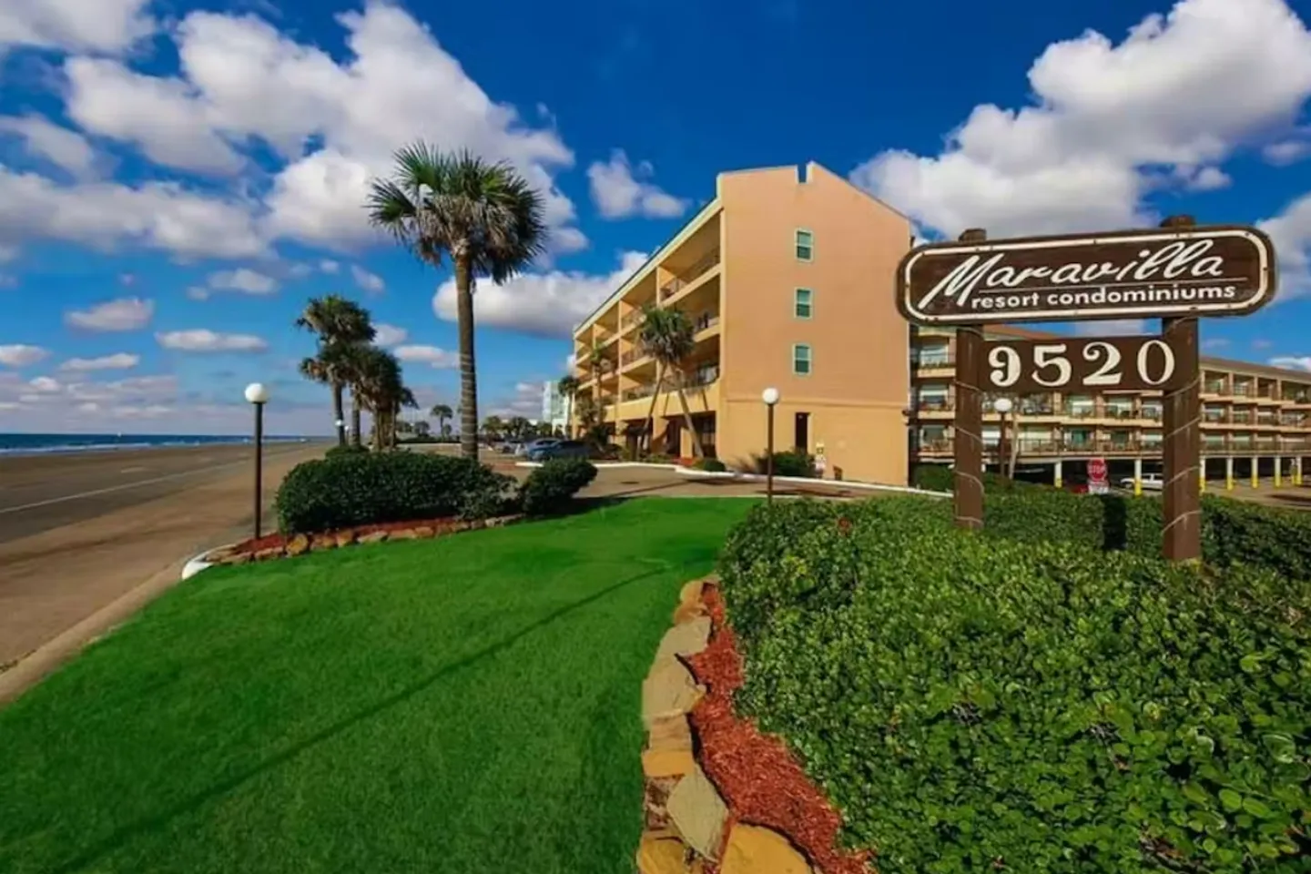 Exterior view of Maravilla Resort Condominiums entrance sign beside a green lawn with the beach and multi-story building in the background.