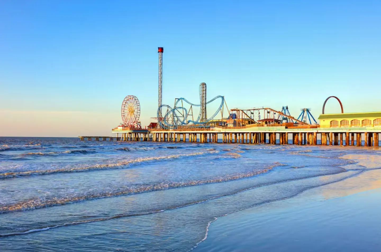 An amusement park pier over the ocean with a roller coaster, Ferris wheel, and drop tower viewed from a sandy beach at sunset.