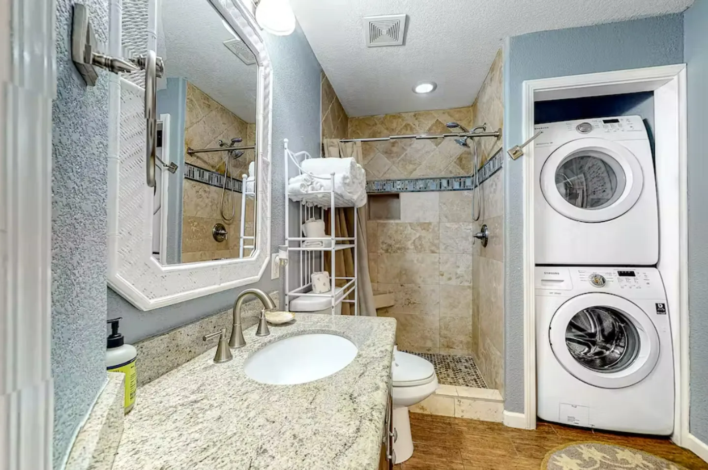 Bathroom with a granite vanity, mirror, toilet, tiled walk-in shower, and a stacked washer and dryer in a closet.