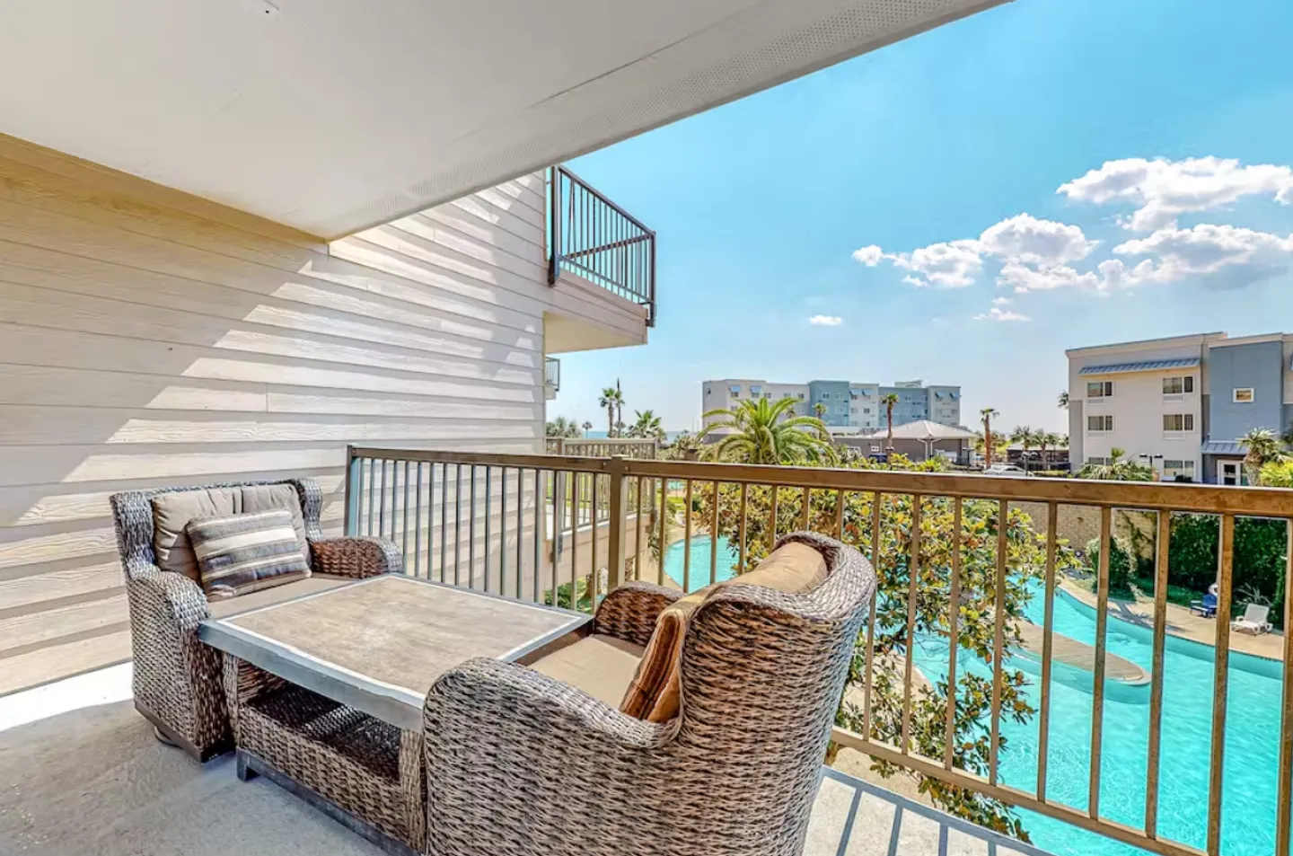 Covered balcony with wicker armchairs and a table overlooking a shared outdoor swimming pool and palm trees under a sunny blue sky.