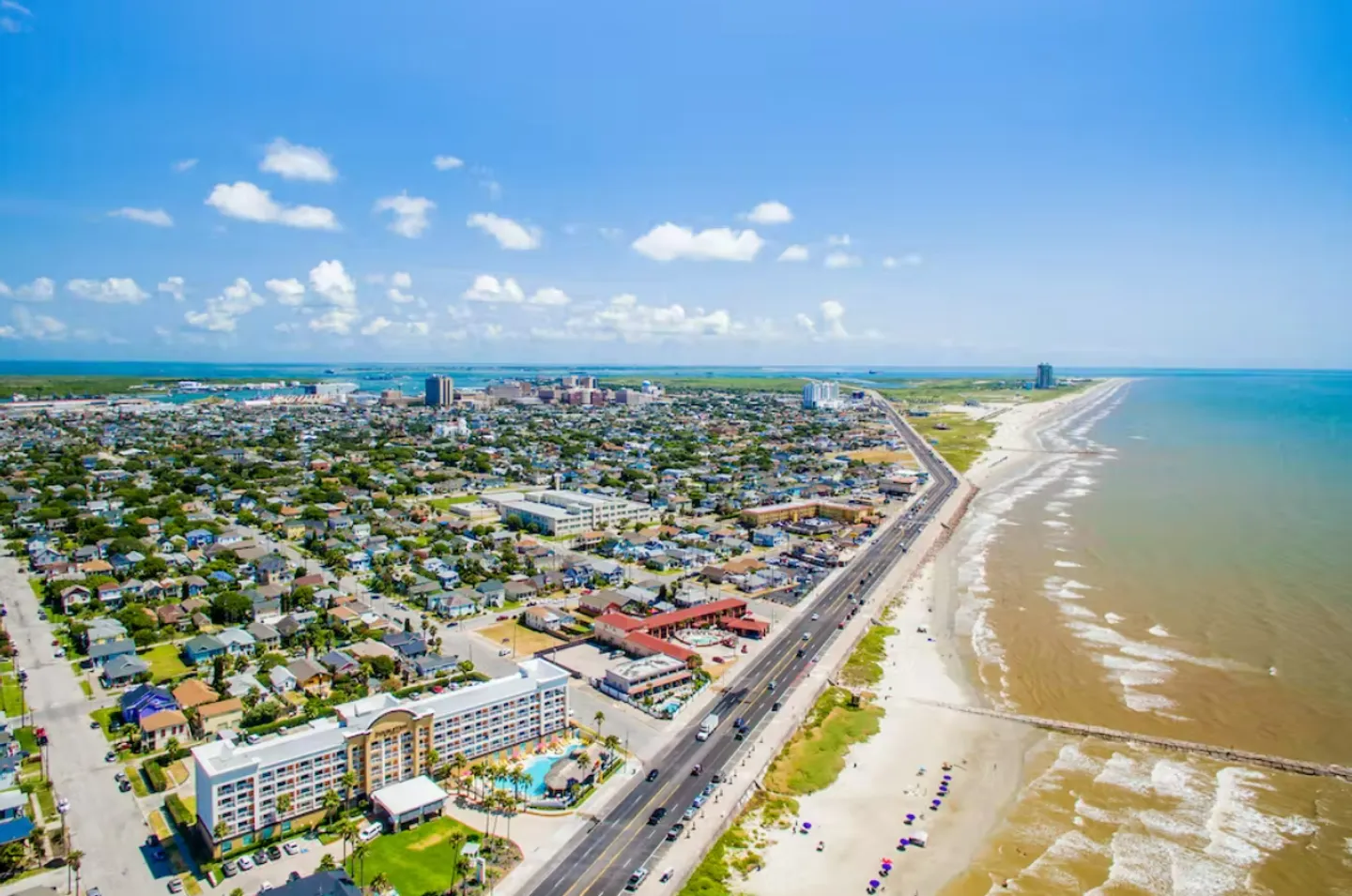 High-angle aerial view of a coastal city, beach, seawall road, and ocean under a blue sky with light clouds.
