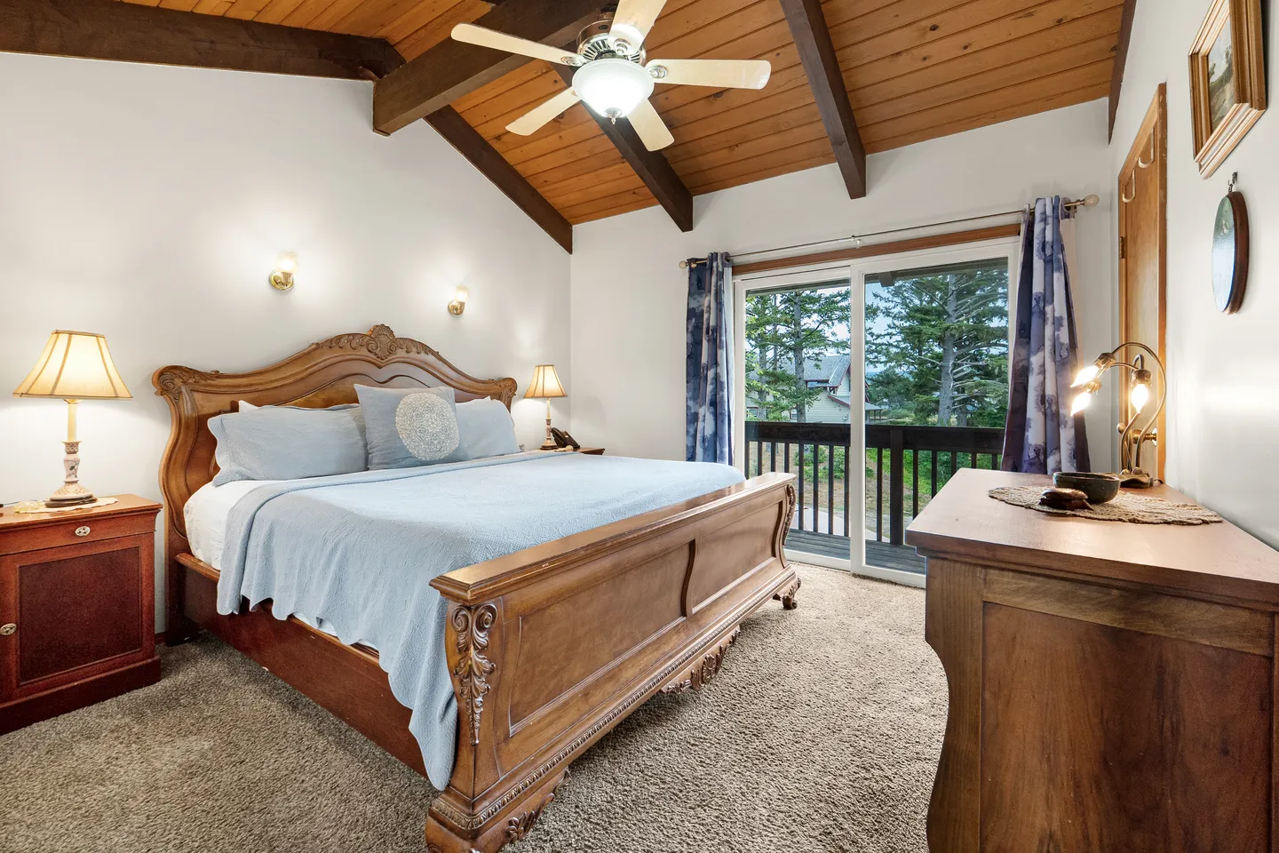 Bedroom featuring an ornate wooden bed frame, vaulted wood-paneled ceiling with exposed beams, and sliding glass doors opening to a balcony with views of trees.