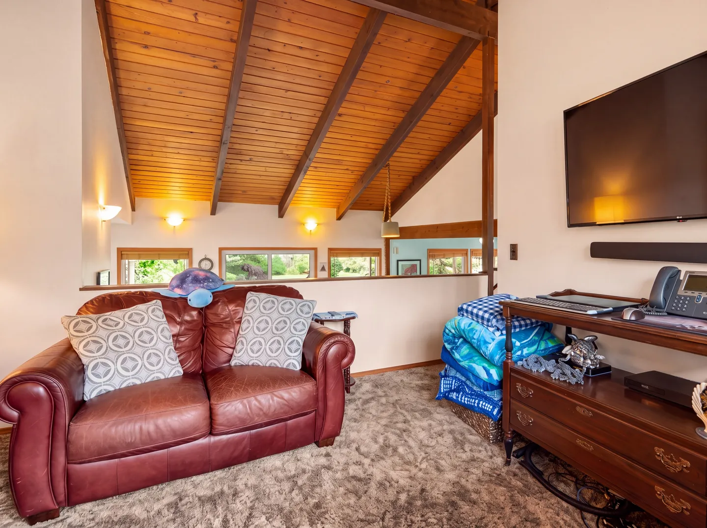 Upstairs loft sitting area with a brown leather loveseat, high vaulted wood-paneled ceiling, and a wall-mounted TV.