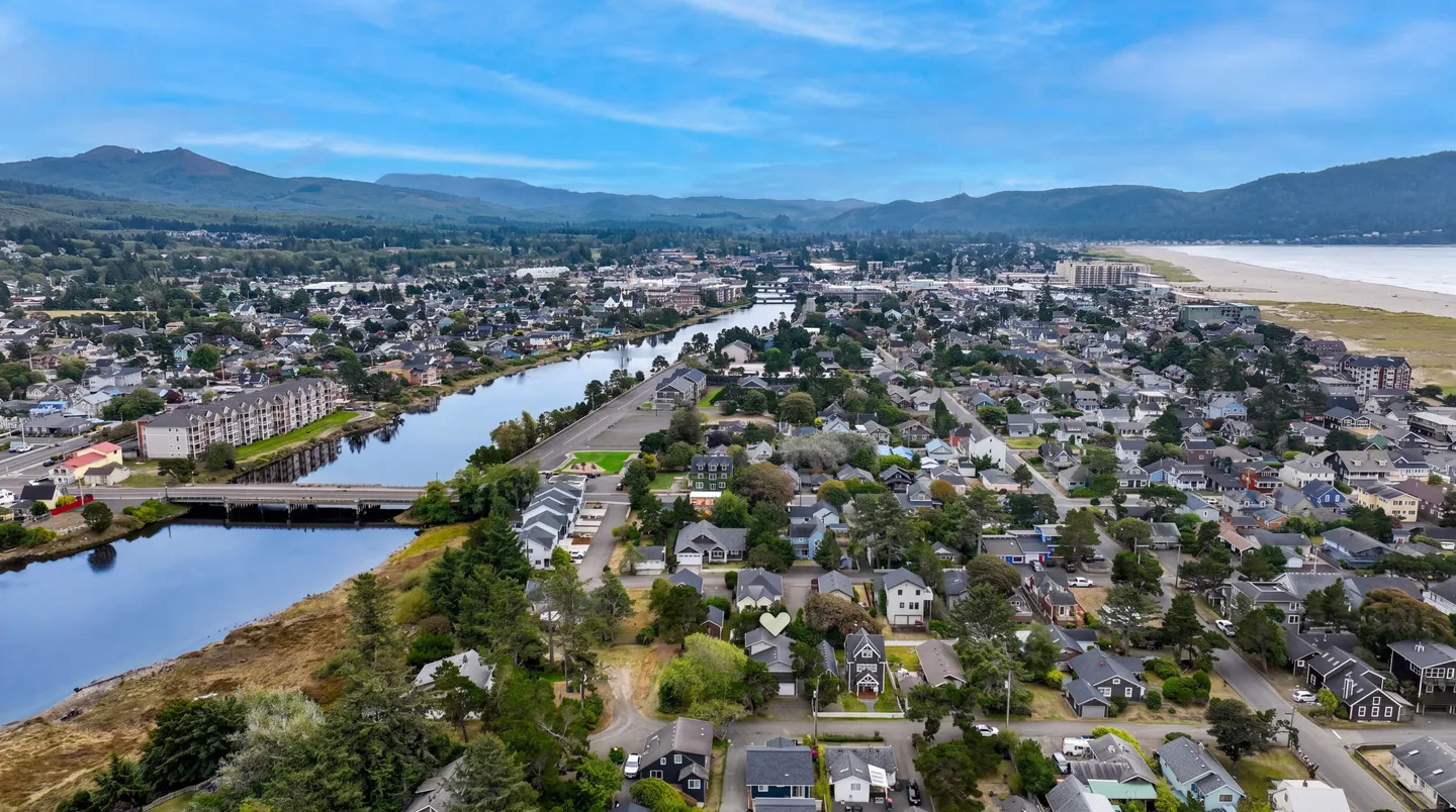 High-angle aerial photograph of a coastal town featuring a river winding through neighborhoods toward the ocean, with forested hills in the distance.