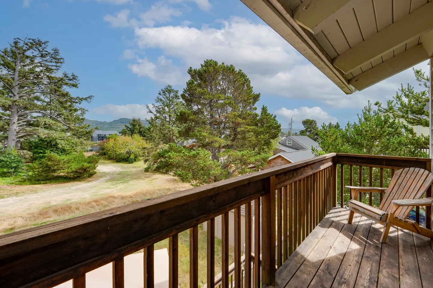 Elevated wooden balcony with an Adirondack chair overlooking green trees and a field under a blue sky.