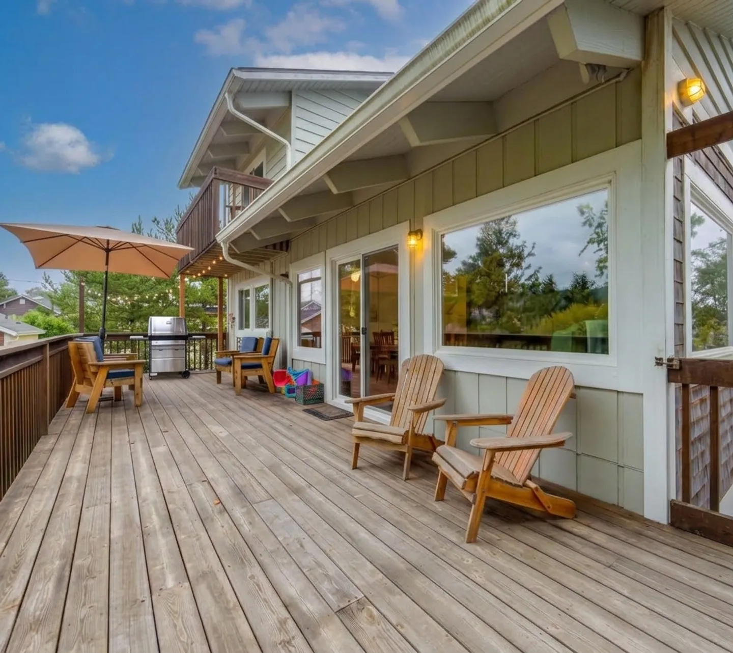 Spacious wooden outdoor deck with Adirondack chairs, an umbrella table set, and a barbecue grill outside a house.