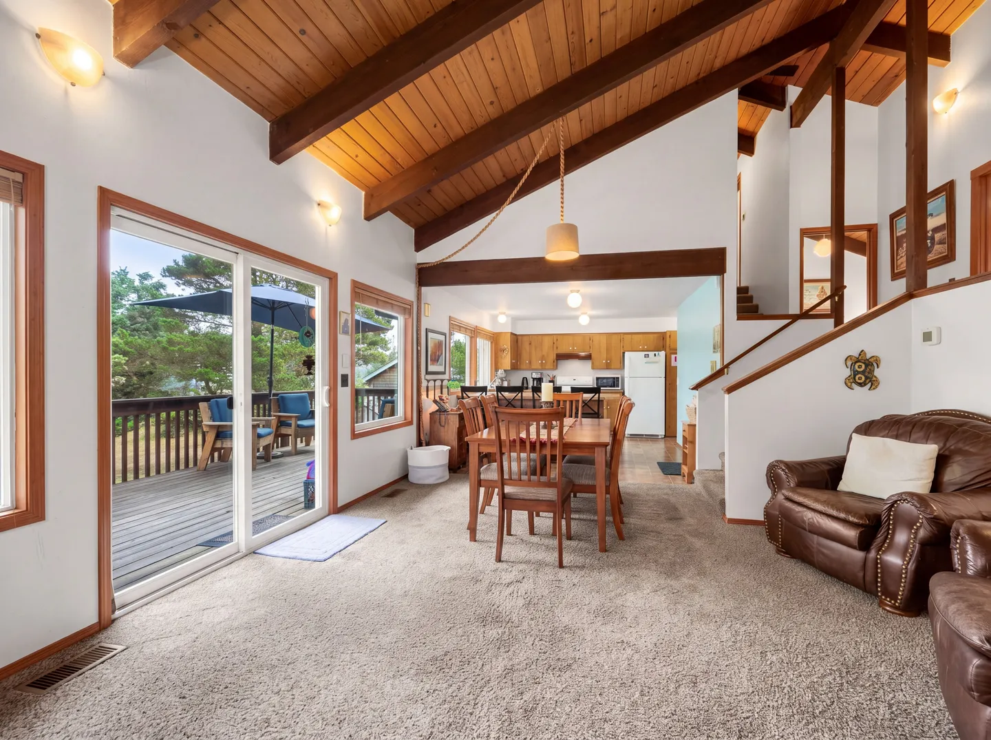 Open living and dining area with carpeted floors, vaulted wood-paneled ceilings with exposed beams, sliding glass doors to a deck, and an adjacent kitchen.