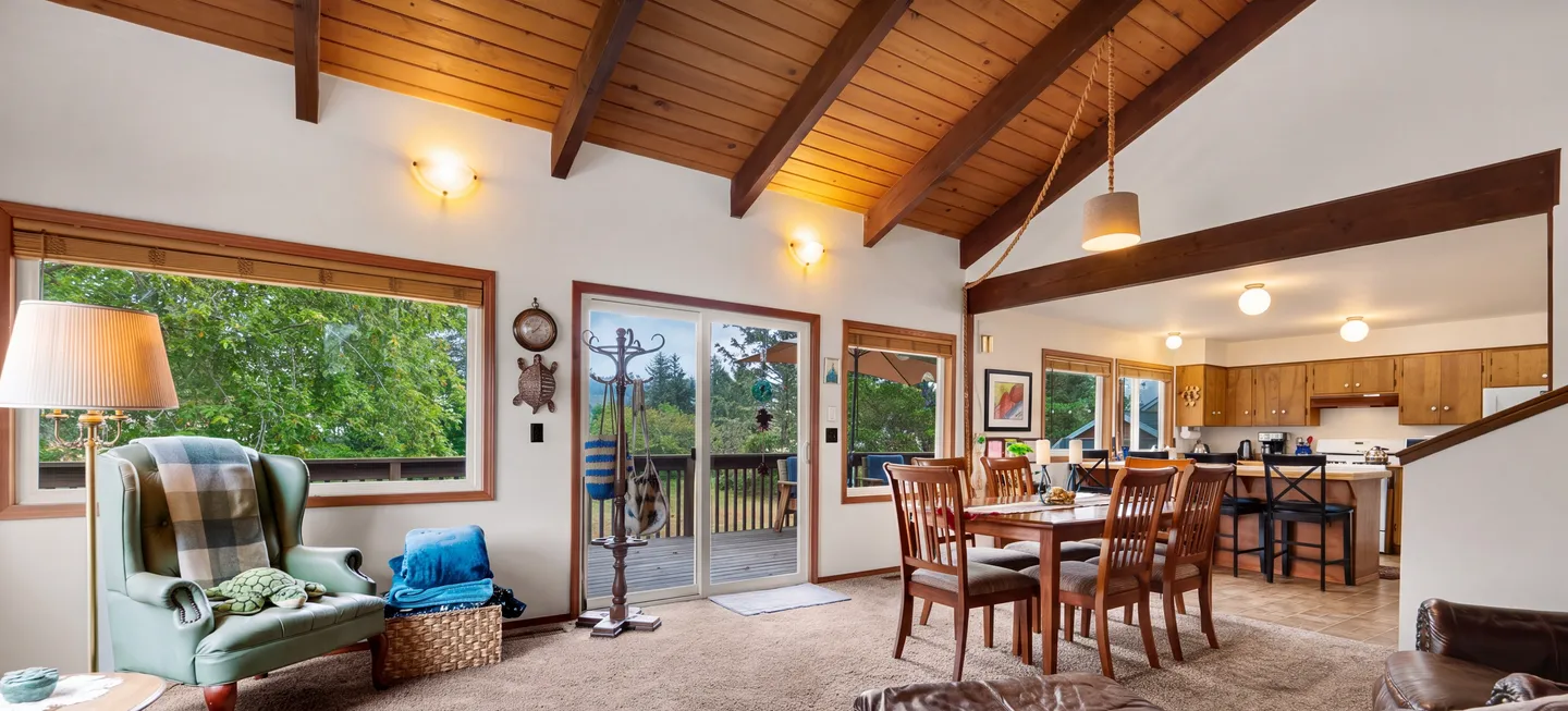 Open-concept living and dining area with vaulted wood-beamed ceilings, an armchair, a dining table with chairs, sliding glass doors to a deck, and an adjacent kitchen.