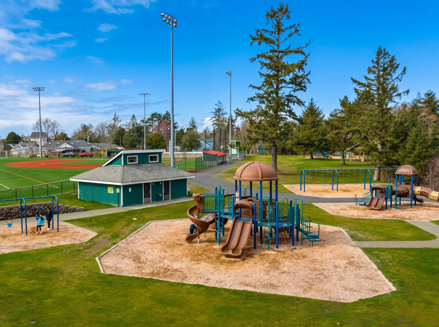Elevated view of a public park playground with play structures, swing sets, a small green building, and a baseball field in the background.