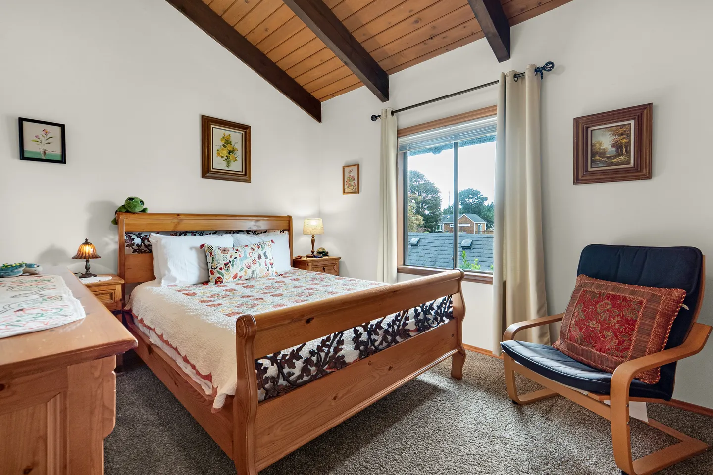 Bedroom featuring a wooden frame bed, vaulted wood-beam ceiling, patterned quilt, nightstand with a lamp, and an armchair next to a window.