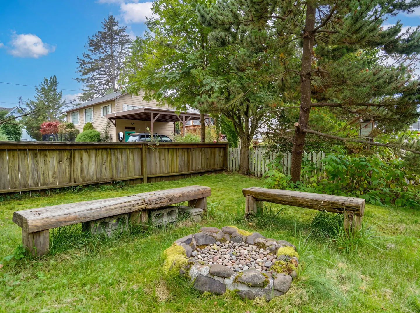 A backyard fire pit made of rocks surrounded by grass and two wooden benches beneath mature trees.