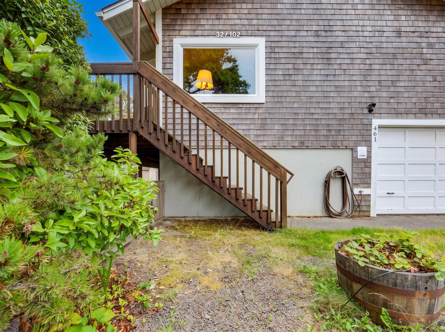 Exterior of a wood-shingled home showing an outdoor wooden staircase leading to an upper deck, a white garage door, and a barrel planter.