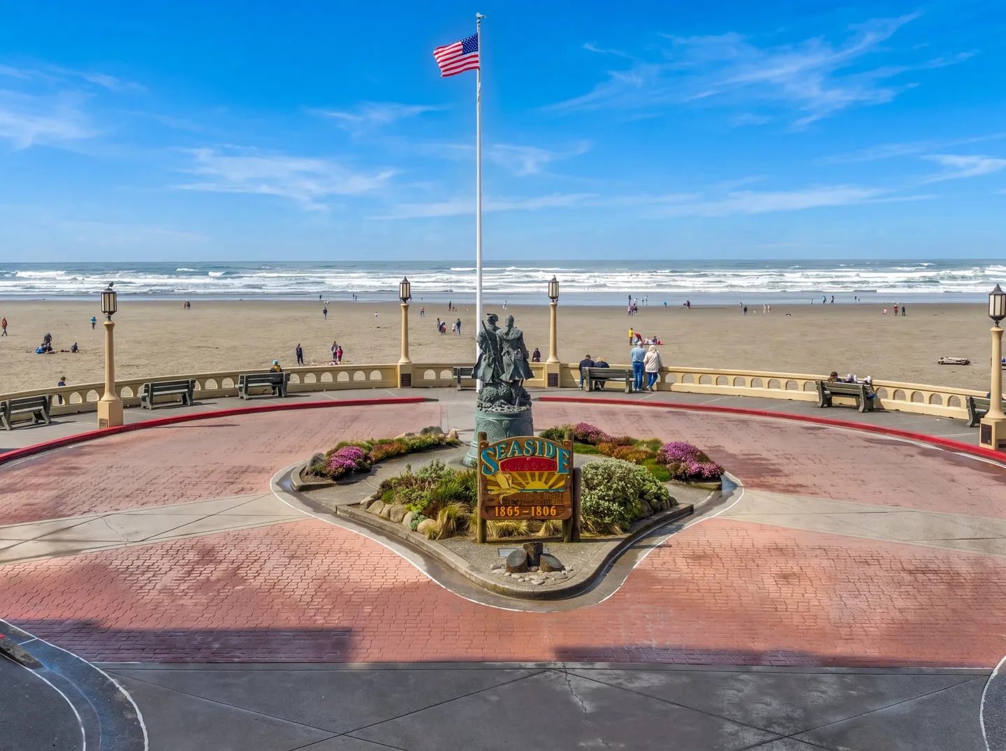 Elevated view of a beach turnaround featuring a flagpole, statue, Seaside sign, and surrounding ocean beach under a blue sky.