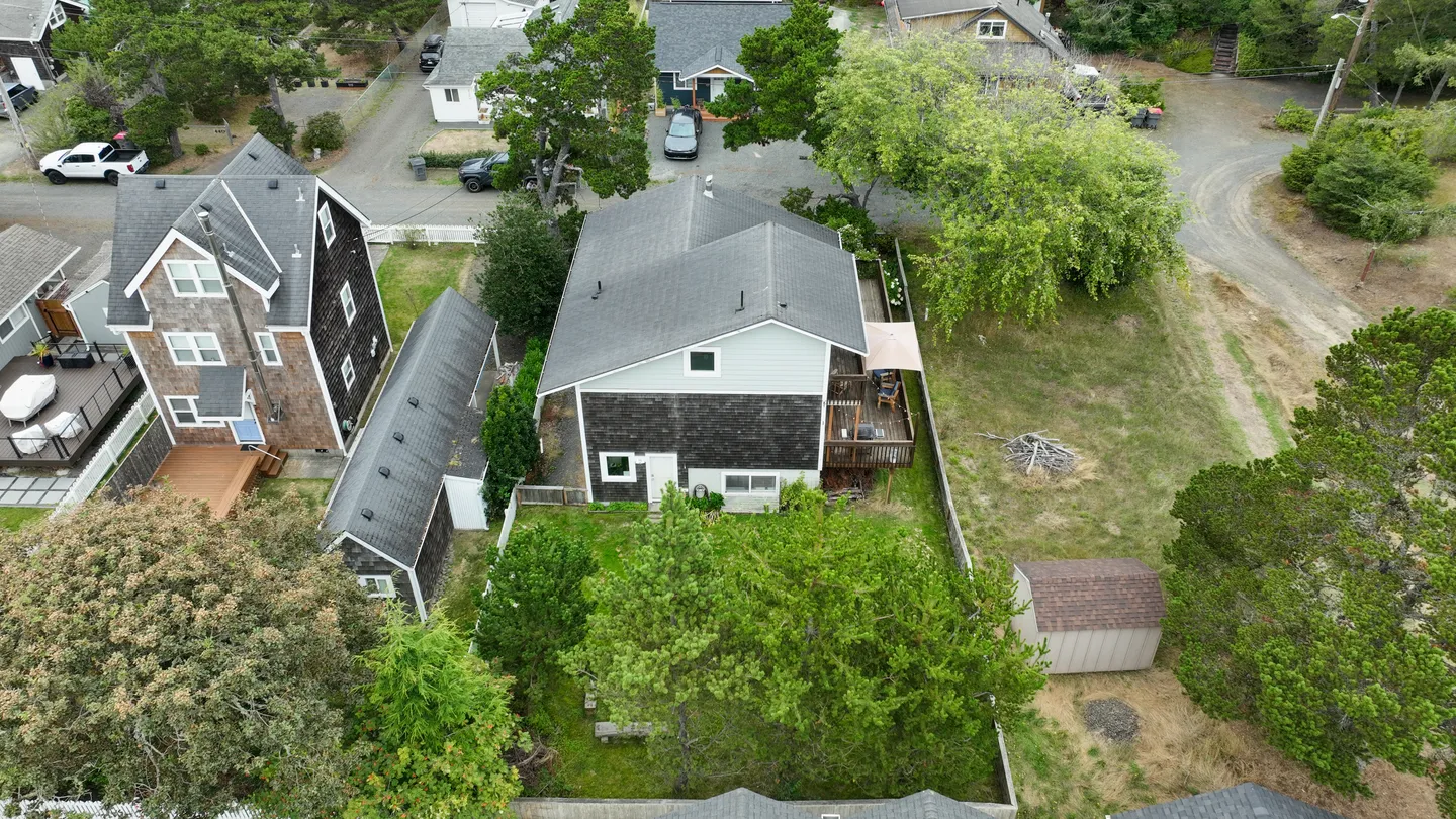 Aerial view of a coastal house with gray shingle and siding exterior, back deck, and surrounding yard bordered by trees.