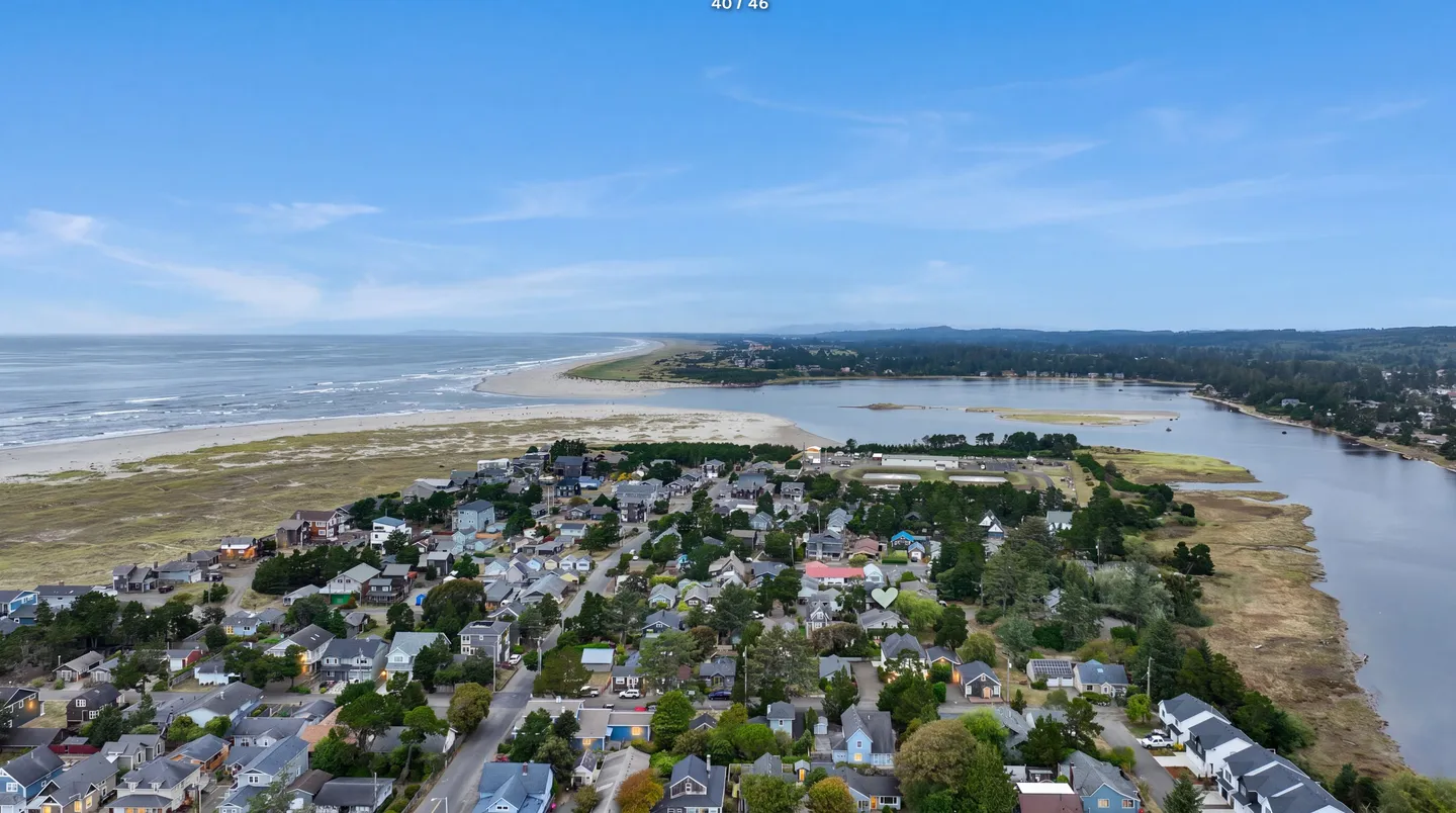 Aerial view of a coastal neighborhood bordered by an ocean beach, a sandy spit, and a calm river under a blue sky.