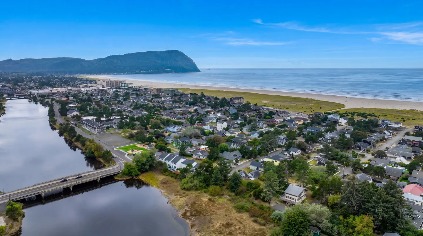 Aerial view of a coastal town bordering a sandy ocean beach, a river crossed by a bridge, and a prominent headland in the background under a blue sky.