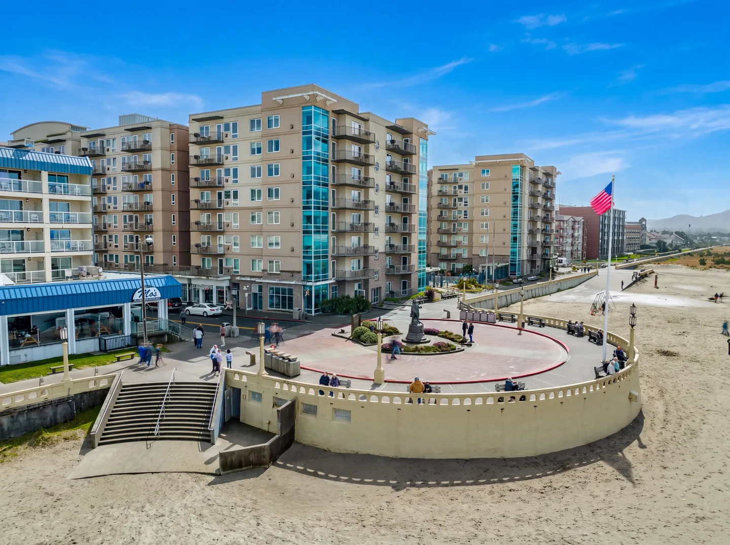 Elevated view of a coastal turnaround plaza adjacent to multi-story buildings and a sandy beach under a clear blue sky.