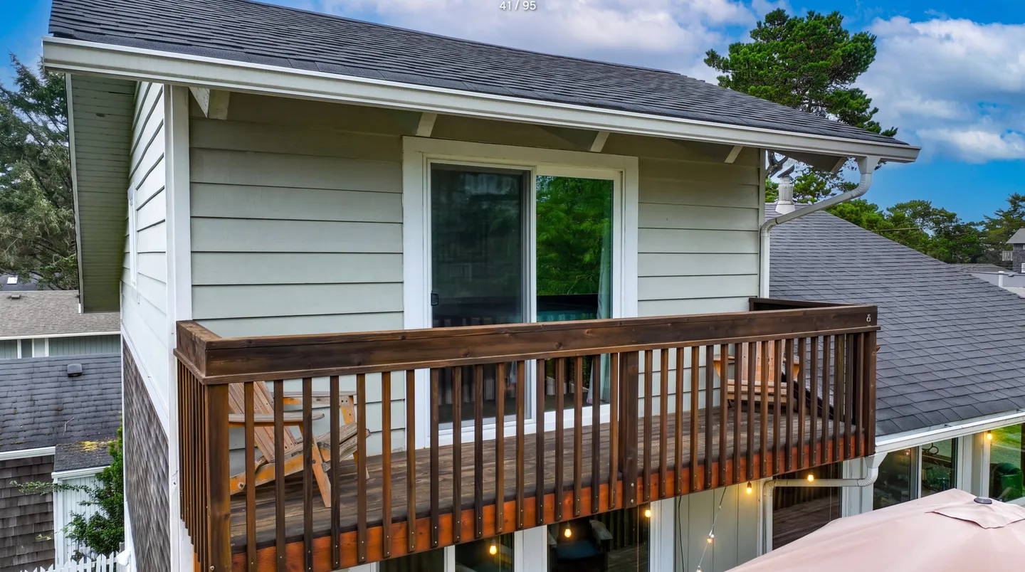 Exterior view of an upper-level wooden balcony with sliding glass doors and outdoor chairs beneath a pitched roof.