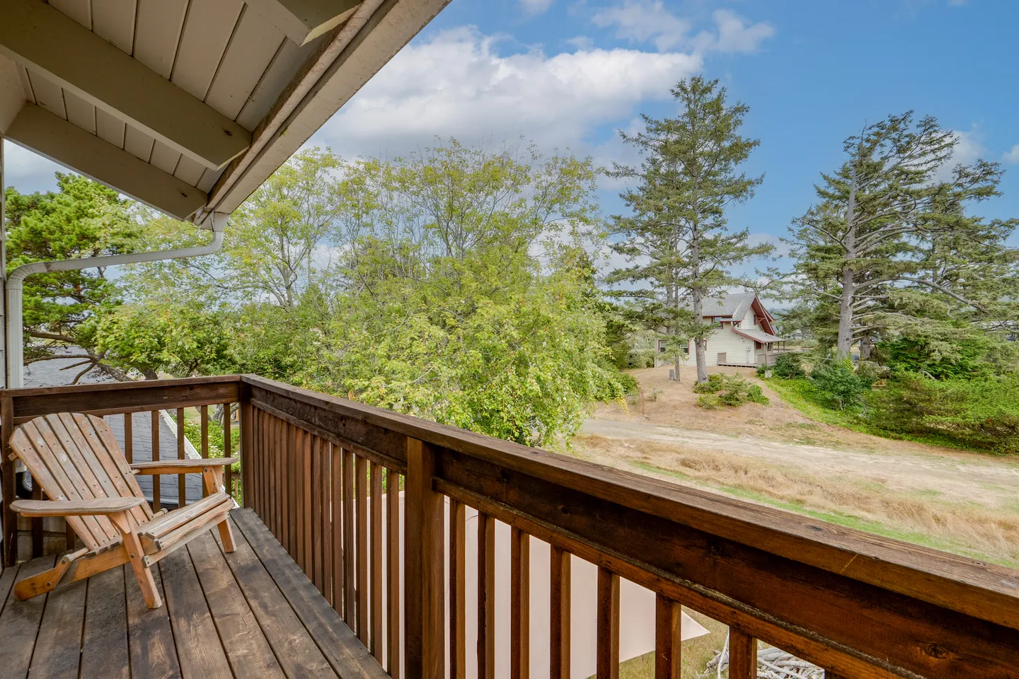 Balcony with a wooden railing and an Adirondack chair overlooking trees and a grassy field with a distant house under a blue sky.