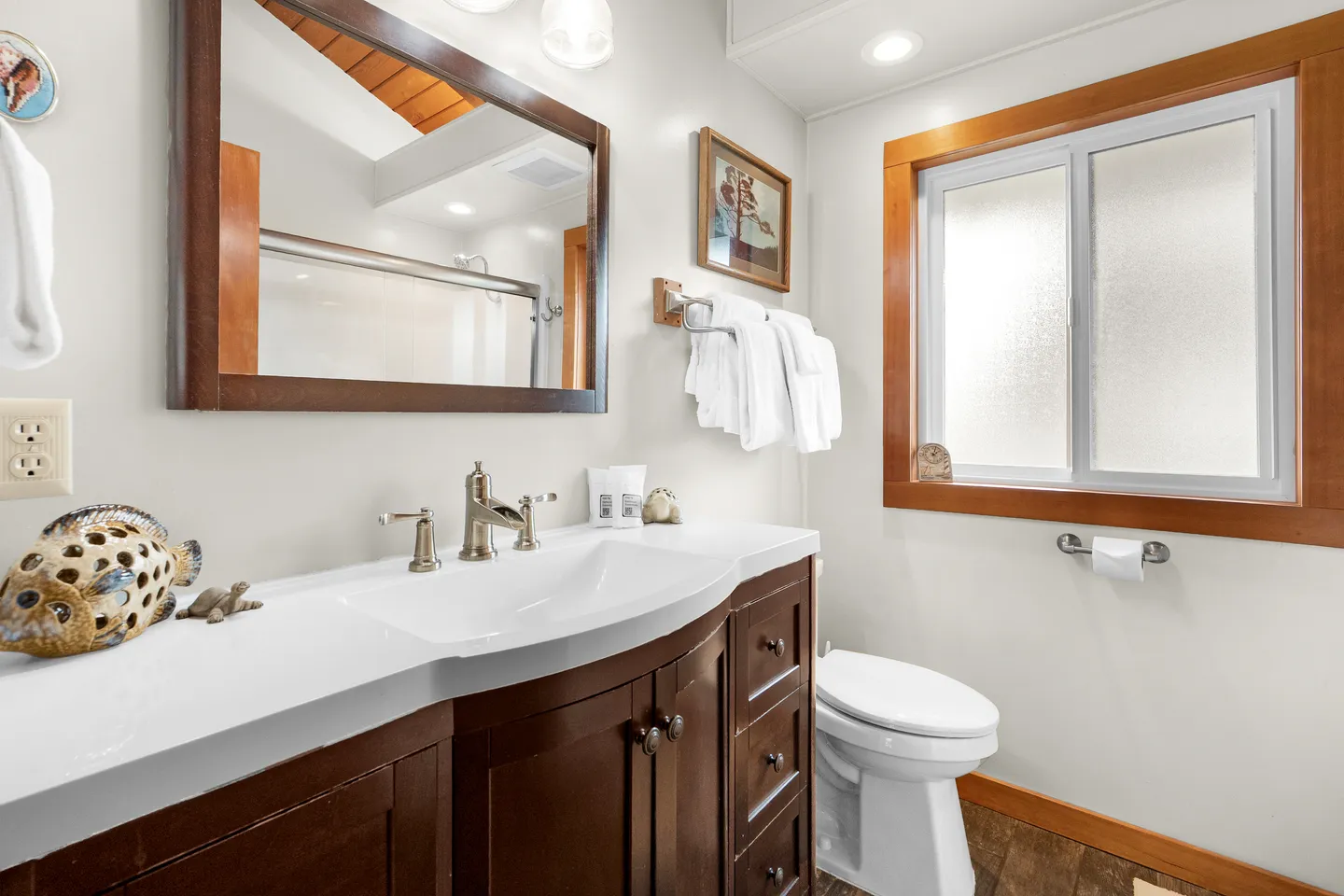 Bathroom with a dark wood vanity, white countertop, large wood-framed mirror, toilet, and frosted glass window.