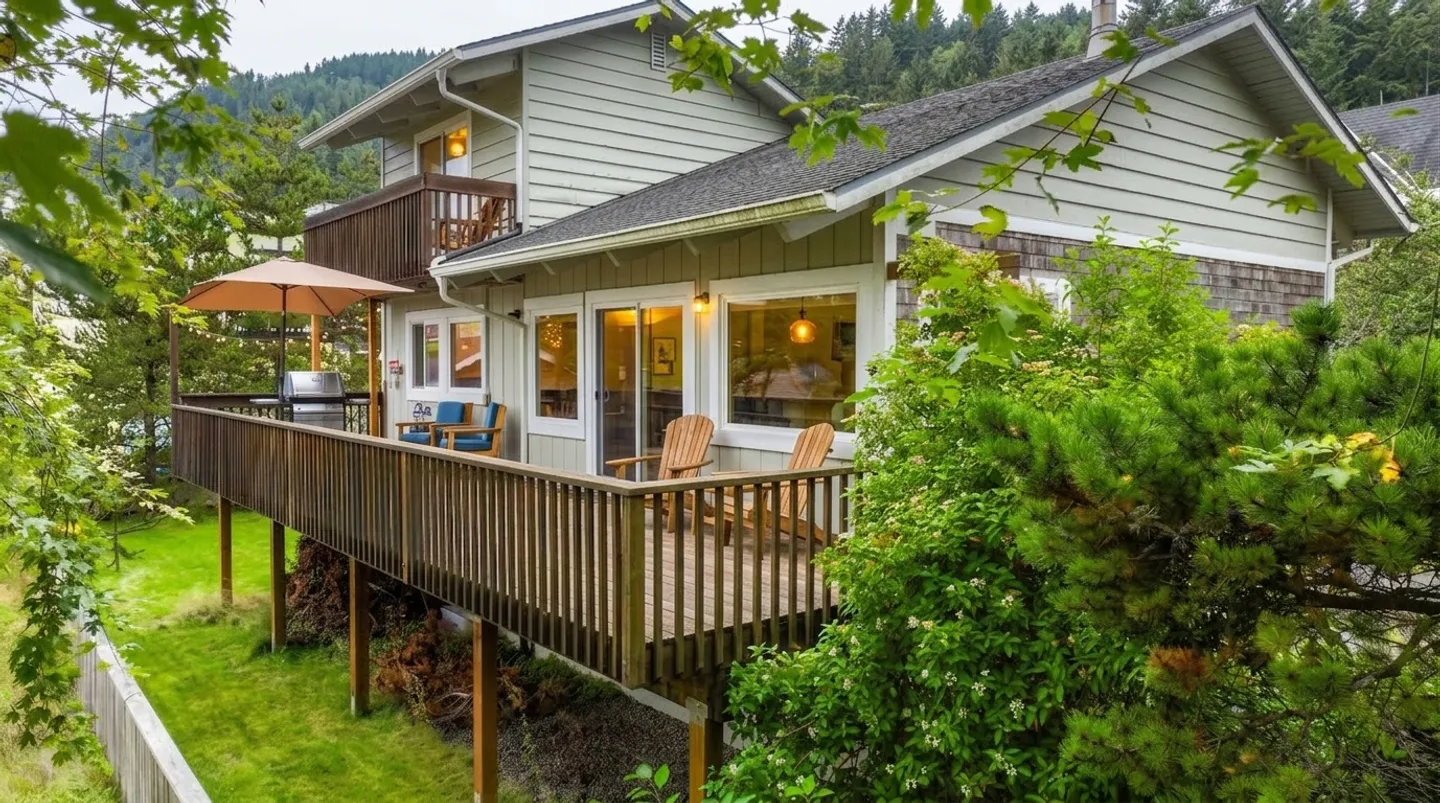 Exterior view of a multi-level green house featuring an elevated wooden deck furnished with chairs, a grill, and an umbrella, surrounded by lush trees.