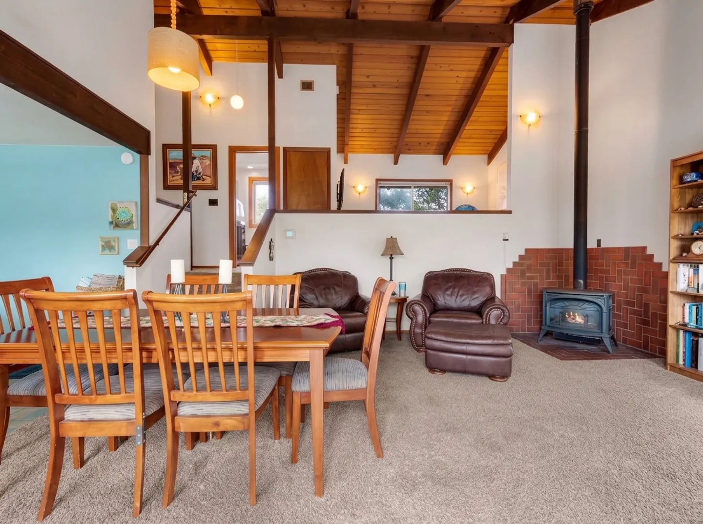 Open-concept dining and living space featuring a wood dining table with chairs, carpeted floor, leather armchairs, a freestanding stove against a brick wall, and wood-paneled vaulted ceilings.