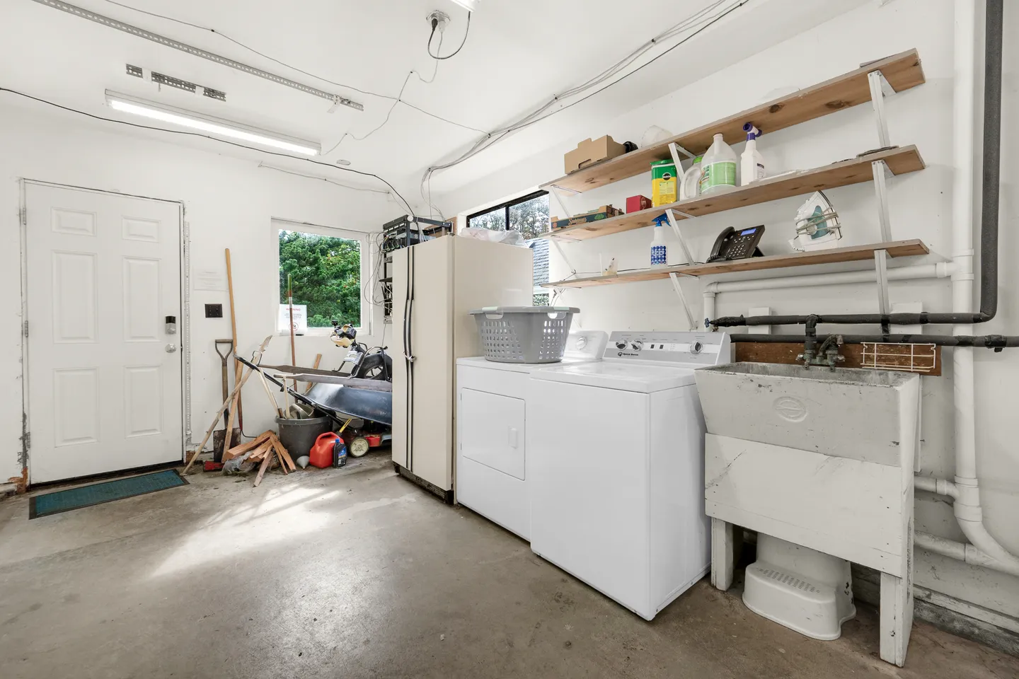 A utility room or garage laundry area featuring a white washer, dryer, utility sink, refrigerator, and open shelving.