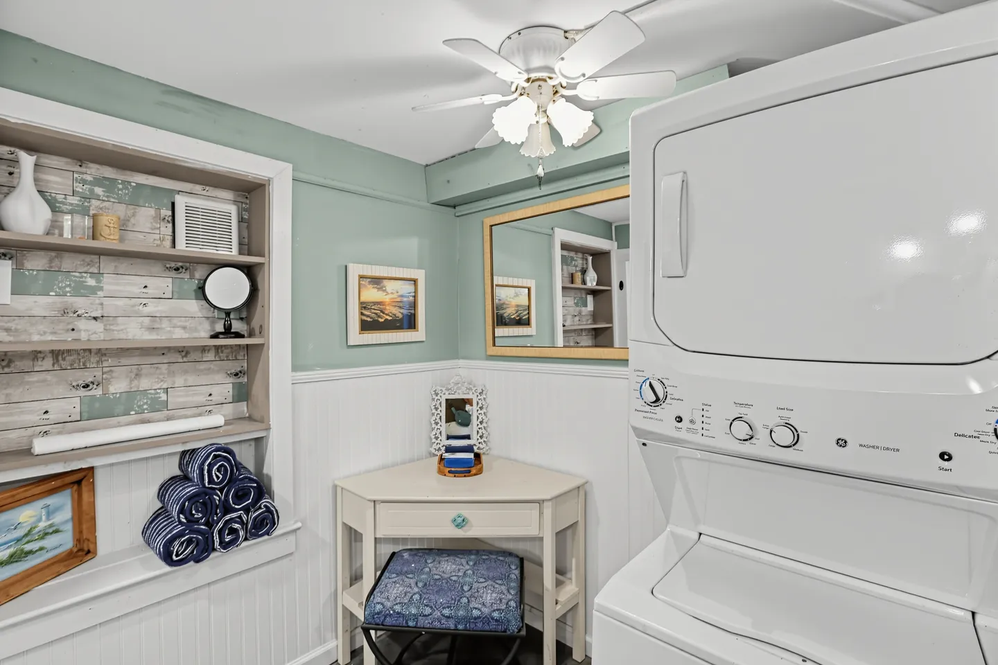 A utility area featuring a stacked washer and dryer, built-in shelving with rolled towels, a corner vanity desk, and a ceiling fan.