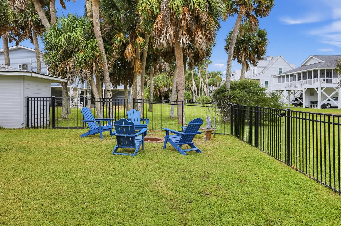 Fenced grassy backyard featuring blue Adirondack chairs encircling a ground fire pit under palm trees.