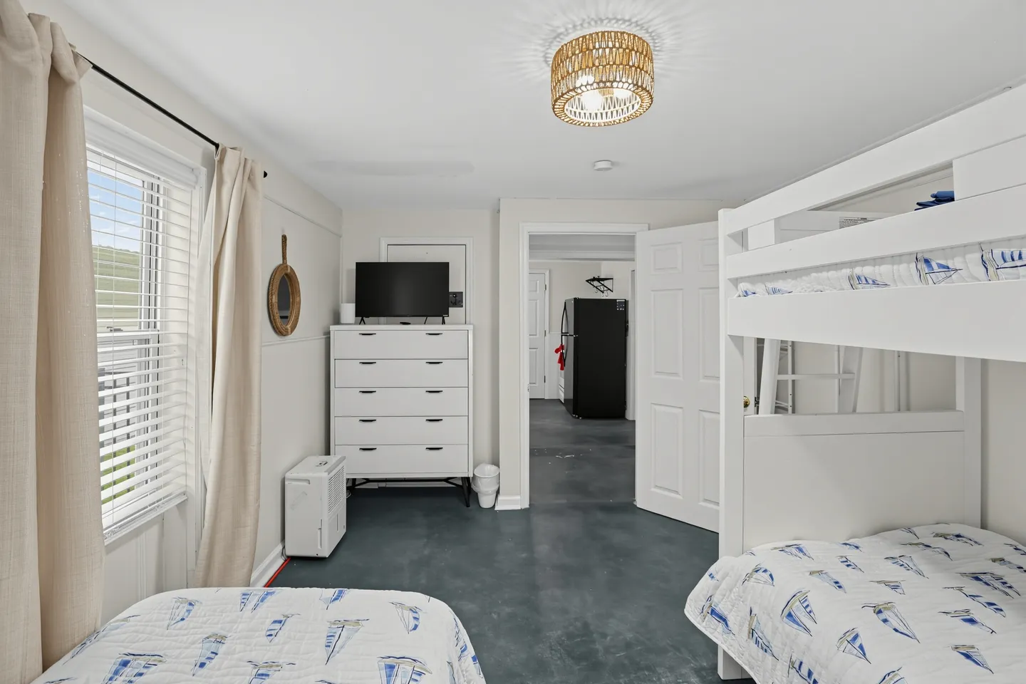 Bedroom featuring white bunk beds with sailboat-patterned bedding, a white dresser with a mounted TV, and an open doorway leading to a kitchen.