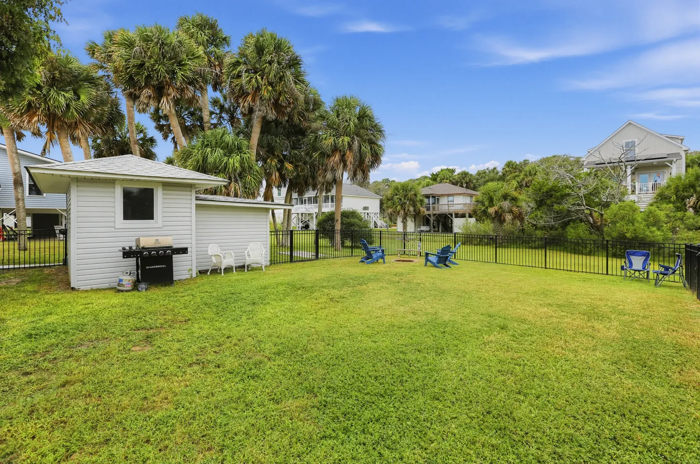 Fenced grassy backyard featuring a fire pit with blue chairs, a small outbuilding with a grill, and palm trees under a blue sky.