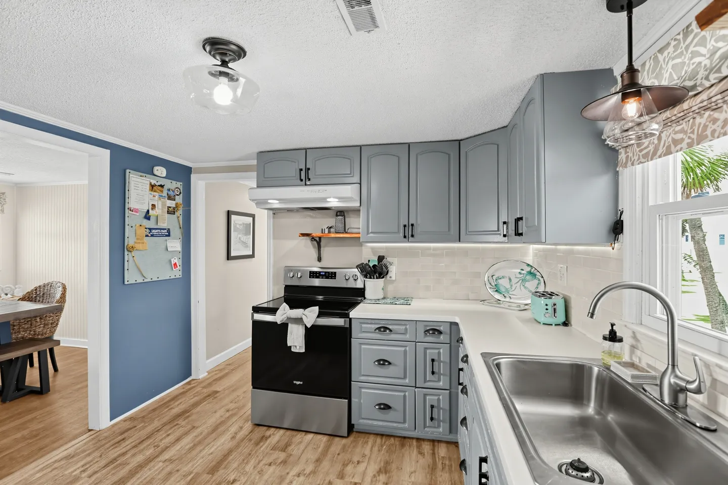 A kitchen with blue-gray cabinets, light countertops, an electric range, and an undermount sink looking into a dining area.