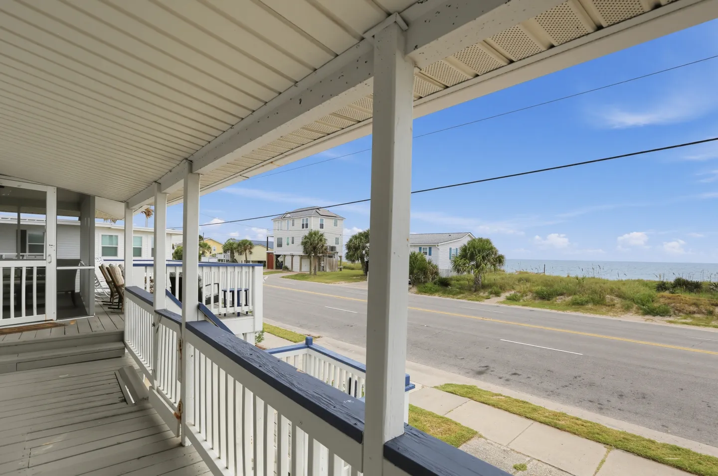 Covered porch with white railings offering a view of a coastal road and the ocean across the street.