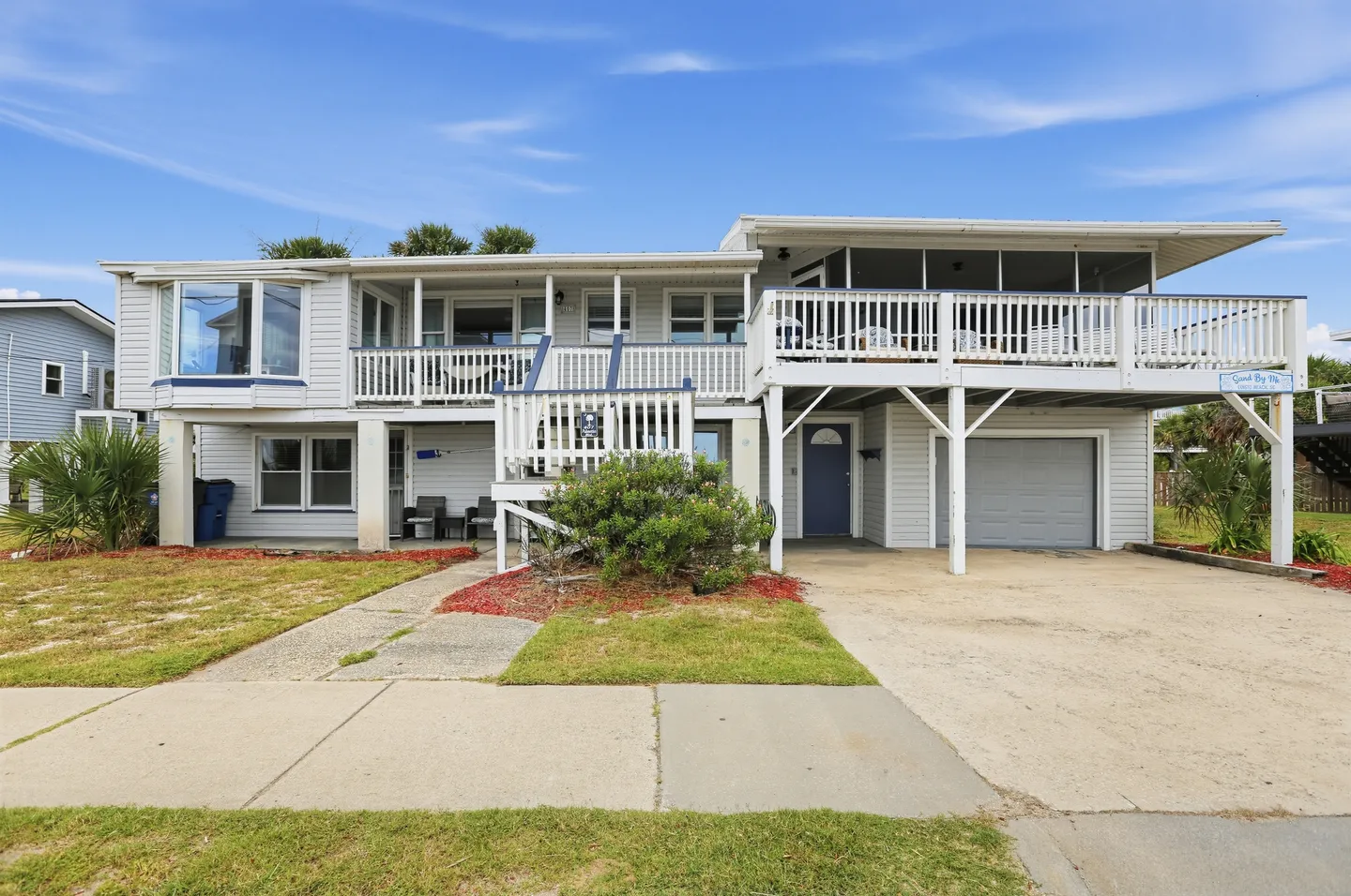 Exterior front view of a two-story coastal beach home with light siding, upper-level deck, screened porch, and driveway.
